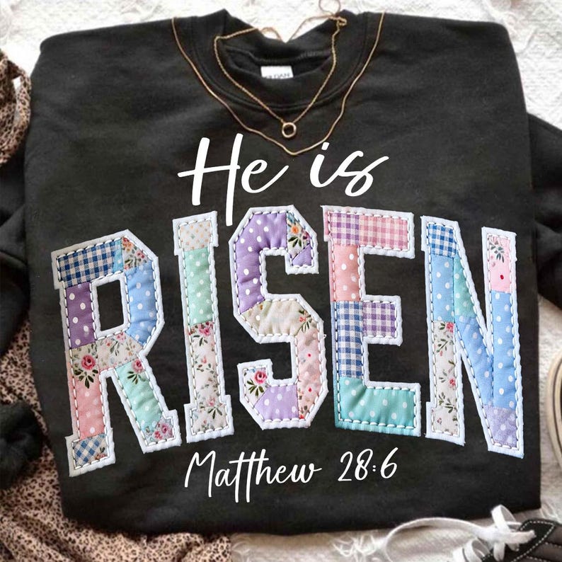 Puede incluir: Sudadera negra con las palabras "He is RISEN" en un dise&ntilde;o de patchwork. Las letras est&aacute;n hechas de varias telas pastel. El texto "Matthew 28:6" est&aacute; debajo de la palabra "RISEN". Un collar dorado est&aacute; drapeado sobre la sudadera.