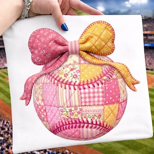 Op de afbeelding: Een decoratief honkbalvormig kussen met een patchwork ontwerp in roze en gele stoffen. Het kussen heeft een grote roze strik bovenop. De honkbal is gemaakt van verschillende stoffen met patronen.