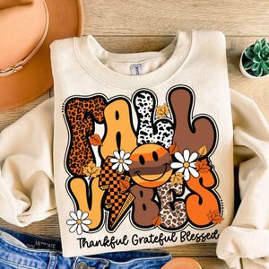 May include: A beige sweatshirt with a graphic design of the words "Fall Vibes" in a colorful, retro style. The design includes a smiley face, flowers, and animal print patterns. The text "Thankful Grateful Blessed" is printed below the graphic.