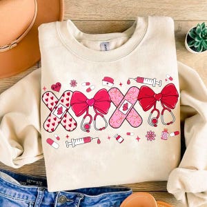 May include: A cream-colored sweatshirt with a graphic design of pink and red band-aids, bows, and medical symbols. The design includes a heart with a heartbeat line, a syringe, and pills.