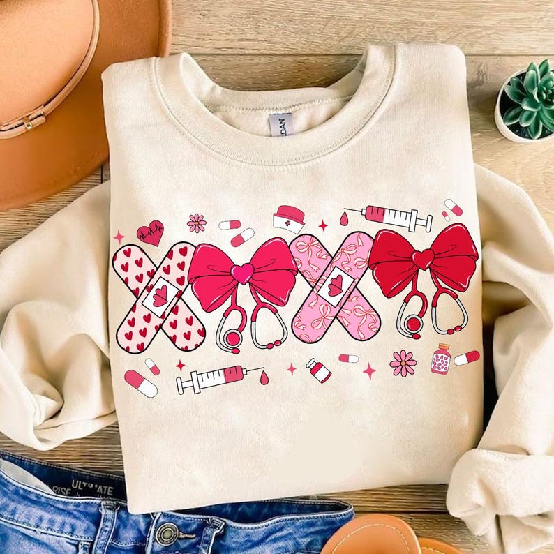 May include: A cream-colored sweatshirt with a graphic design of pink and red band-aids, bows, and medical symbols. The design includes a heart with a heartbeat line, a syringe, and pills.