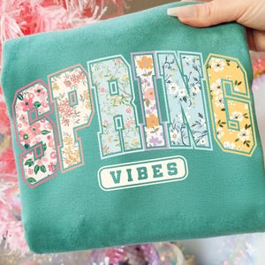 May include: Teal sweatshirt with the word "SPRING" in a floral design, and the word "VIBES" in a white, rounded rectangle. The letters are filled with various floral patterns in pink, yellow, and blue.