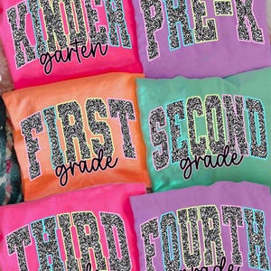 May include: A collection of colorful t-shirts, each displaying a grade level. The shirts are in shades of pink, orange, purple, and green. The text, outlined in blue and yellow, is filled with a glittery pattern. The shirts are labeled "Kindergarten", "Pre-K", "First Grade", "Second Grade", "Third Grade", and "Fourth Grade".