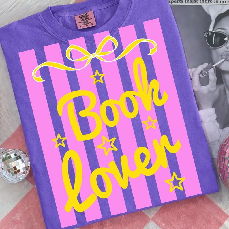 May include: A purple t-shirt featuring a pink striped design with the words "Book Lover" in yellow. The design includes a yellow bow and stars. The shirt is made of a soft material.
