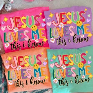 May include: Four colorful t-shirts with the text "JESUS LOVES ME this i know" in a playful, multi-colored design. Each shirt is a different color: pink, purple, orange, and teal. The design includes smiley faces, hearts, and stars.