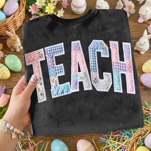 Może przedstawiać: Czarna bluza z napisem "TEACH" z aplikacją z patchworku. Litery są obramowane na biało i mają wzory kwiatowe, w kratkę i jednolite pastele. Bluza jest prezentowana na drewnianej powierzchni z dekoracjami wielkanocnymi.