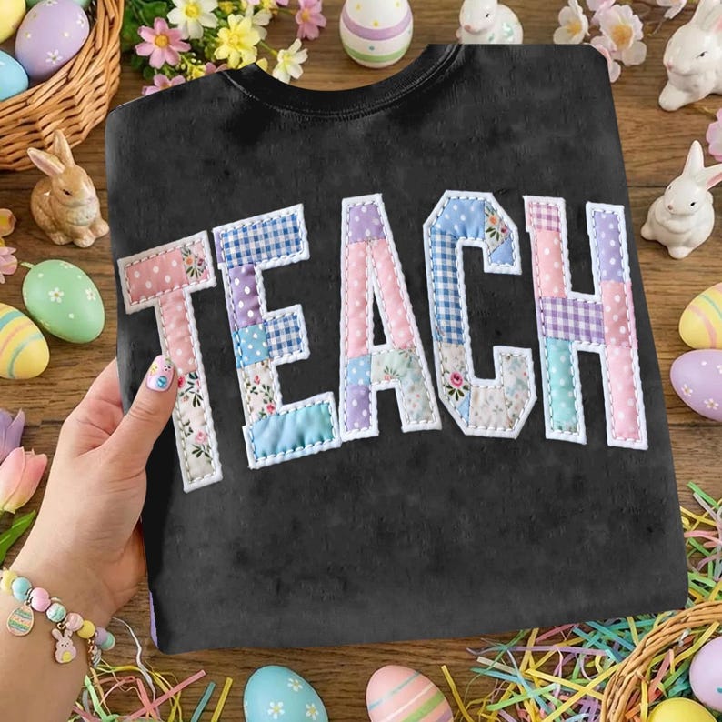 Może przedstawiać: Czarna bluza z napisem "TEACH" z aplikacją z patchworku. Litery są obramowane na biało i mają wzory kwiatowe, w kratkę i jednolite pastele. Bluza jest prezentowana na drewnianej powierzchni z dekoracjami wielkanocnymi.