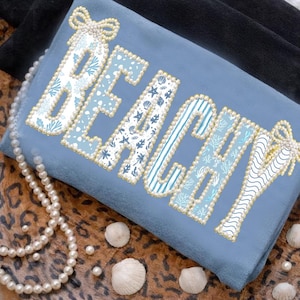 Puede incluir: Un cojín decorativo azul claro con la palabra "BEACHY" bordada en patrones blancos y azules, delineada con perlas de imitación. La imagen incluye conchas marinas y un collar de perlas.