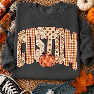 May include: A dark gray crewneck sweatshirt featuring the word CUSTOM in large block letters. The letters are filled with various patterns including orange and white gingham, floral prints, and brown polka dots, with an orange pumpkin graphic.