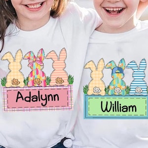 Puede incluir: Dos camisetas blancas con un diseño de tres conejos de Pascua en fila. Los conejos llevan ropa de diferentes colores y tienen diferentes patrones. Las camisetas están personalizadas con los nombres "Adalynn" y "William".
