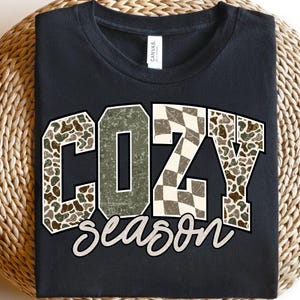K&ouml;nnte beinhalten: Schwarzes T-Shirt mit dem Schriftzug "COZY season" in dekorativer Schrift. Die Buchstaben "C" und "Y" sind mit einem Camouflage-Muster gef&uuml;llt, w&auml;hrend das "Z" ein Schachbrettmuster aufweist. Das T-Shirt wird auf einer gewebten Oberfl&auml;che pr&auml;sentiert.