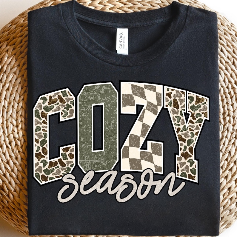 K&ouml;nnte beinhalten: Schwarzes T-Shirt mit dem Schriftzug "COZY season" in dekorativer Schrift. Die Buchstaben "C" und "Y" sind mit einem Camouflage-Muster gef&uuml;llt, w&auml;hrend das "Z" ein Schachbrettmuster aufweist. Das T-Shirt wird auf einer gewebten Oberfl&auml;che pr&auml;sentiert.