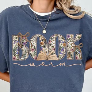K&ouml;nnte beinhalten: Ein marineblaues Sweatshirt mit einem floralen Design, das "BOOK WORM" buchstabiert. Das Wort "BOOK" steht in gro&szlig;en Buchstaben mit einem offenen Buch in der Mitte. Das Wort "WORM" ist in Kursivschrift unter dem Wort "BOOK" geschrieben.
