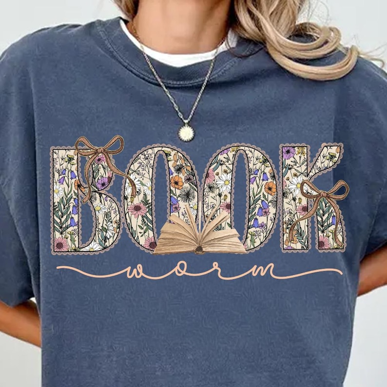 K&ouml;nnte beinhalten: Ein marineblaues Sweatshirt mit einem floralen Design, das "BOOK WORM" buchstabiert. Das Wort "BOOK" steht in gro&szlig;en Buchstaben mit einem offenen Buch in der Mitte. Das Wort "WORM" ist in Kursivschrift unter dem Wort "BOOK" geschrieben.