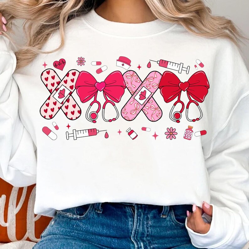 May include: White sweatshirt with a pink and red graphic design of bandaged hearts, bows, and stethoscopes. The graphic includes the text "XXX".