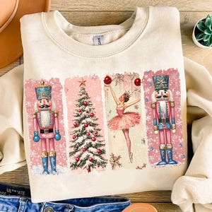 May include: A cream coloured sweatshirt with a pink and white Christmas design. The design features a ballerina, a Christmas tree, and two nutcracker soldiers.