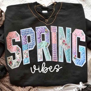 May include: Black sweatshirt with the word "SPRING" in patchwork letters, featuring floral and polka dot patterns. The word "vibes" is written below in cursive. A gold necklace rests on the sweatshirt.