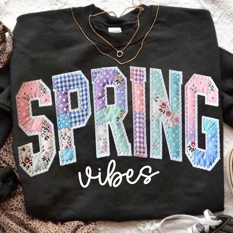 May include: Black sweatshirt with the word "SPRING" in patchwork letters, featuring floral and polka dot patterns. The word "vibes" is written below in cursive. A gold necklace rests on the sweatshirt.