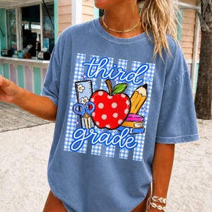 K&ouml;nnte beinhalten: Hellblaues T-Shirt mit einem Aufdruck "third grade". Das Design zeigt einen roten Apfel mit wei&szlig;en Punkten, einen Bleistift, eine Schere und ein Lineal, vor einem blau-wei&szlig; karierten Hintergrund.