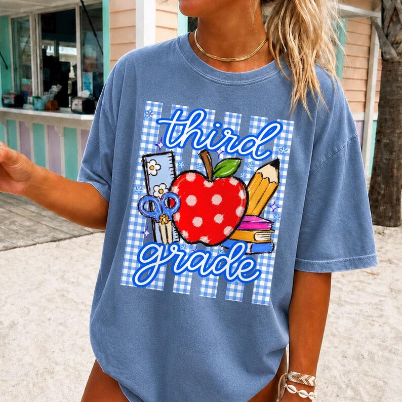 K&ouml;nnte beinhalten: Hellblaues T-Shirt mit einem Aufdruck "third grade". Das Design zeigt einen roten Apfel mit wei&szlig;en Punkten, einen Bleistift, eine Schere und ein Lineal, vor einem blau-wei&szlig; karierten Hintergrund.