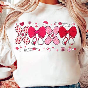 May include: White sweatshirt with a pink and red design featuring a stethoscope, bandages, and a syringe. The design is centered on the front of the sweatshirt.