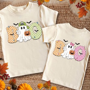 Könnte beinhalten: Zwei weiße T-Shirts mit dem Wort "BOO" in bunten Karomustern. Jeder Buchstabe hat einen Geist, der einen Kürbis hält.