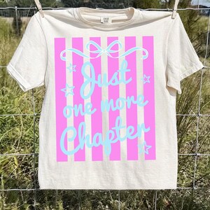 May include: A cream-coloured t-shirt featuring a pink striped design with the text "Just one more chapter" in light blue script. The design includes a bow and stars. The t-shirt is hanging on a clothesline.