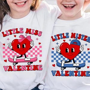 May include: Two white t-shirts with a Valentine's Day theme. The shirt on the left says "Little Miss Valentine" and features a red heart with a pink bow and a skateboard. The shirt on the right says "Little Mister Valentine" and features a red heart with a blue hat and a skateboard.