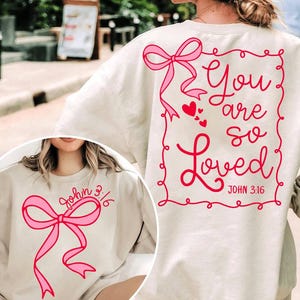 May include: Two cream-colored sweatshirts with pink bows and the text "You are so Loved" and "John 3:16" in red.