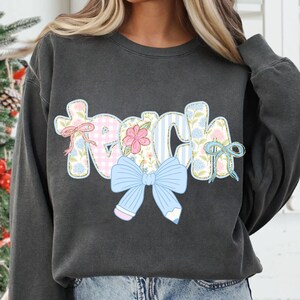 Puede incluir: Una sudadera gris oscura con la palabra "Teach" en letras de patchwork de colores con un lazo azul y detalles florales.