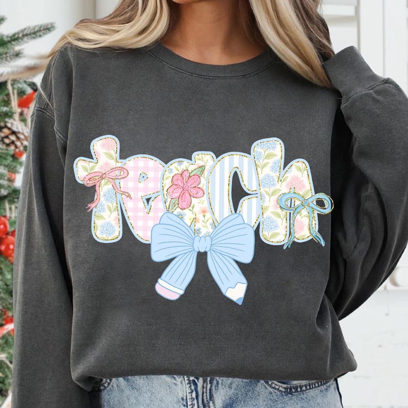 Puede incluir: Una sudadera gris oscura con la palabra "Teach" en letras de patchwork de colores con un lazo azul y detalles florales.