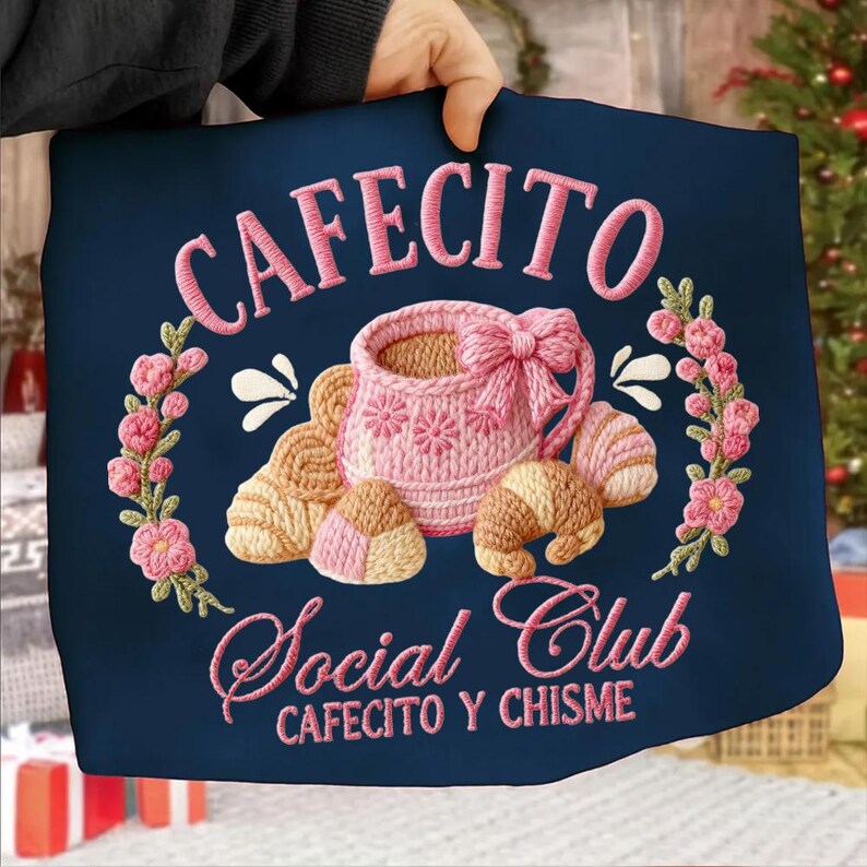 Pode incluir: Saco tote azul marinho com design bordado. Apresenta o texto "CAFECITO Social Club" e uma ch&aacute;vena de caf&eacute; rosa com um la&ccedil;o, rodeada por croissants e detalhes florais. O design &eacute; em tons de rosa e bege.
