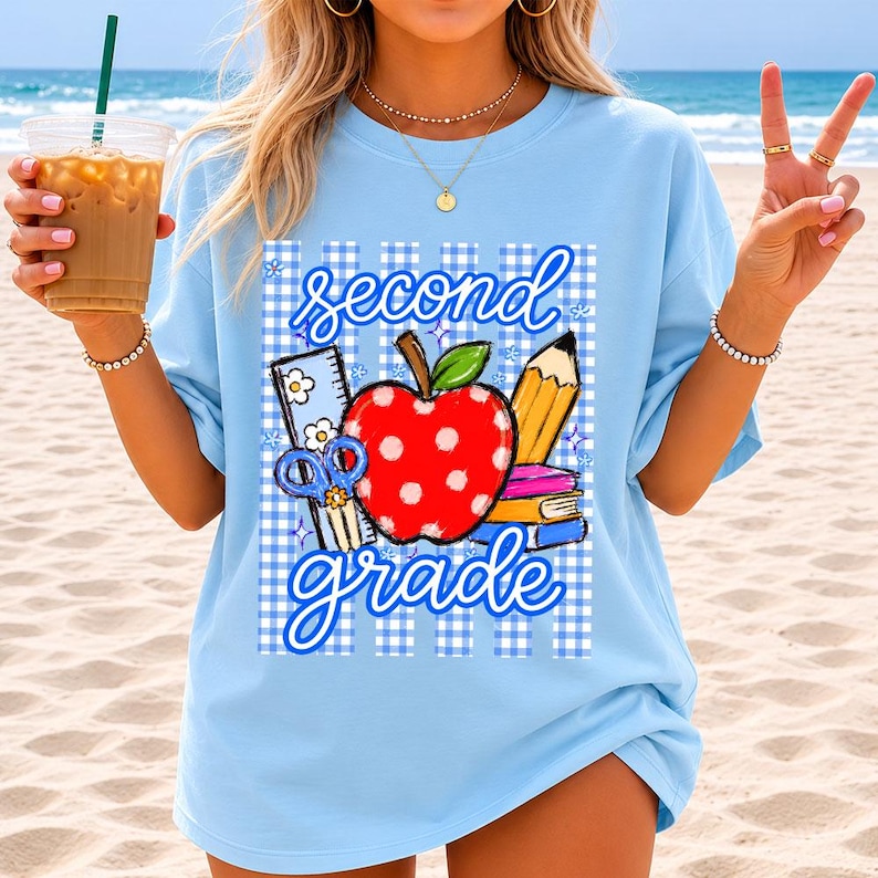 K&ouml;nnte beinhalten: Hellblaues T-Shirt mit einem Grafikdesign. Das Design zeigt einen roten Apfel mit wei&szlig;en Punkten, einen gelben Bleistift und die Worte "second grade". Das Design umfasst auch Schulmaterial und ein blau-wei&szlig;es Karomuster.