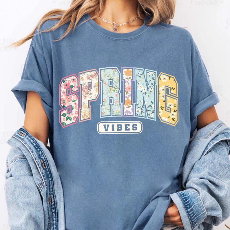 Puede incluir: Camiseta azul con la frase "SPRING VIBES". La palabra "SPRING" est&aacute; en una fuente de estilo universitario con estampados florales en rosa, azul y amarillo. "VIBES" est&aacute; en un rect&aacute;ngulo blanco.