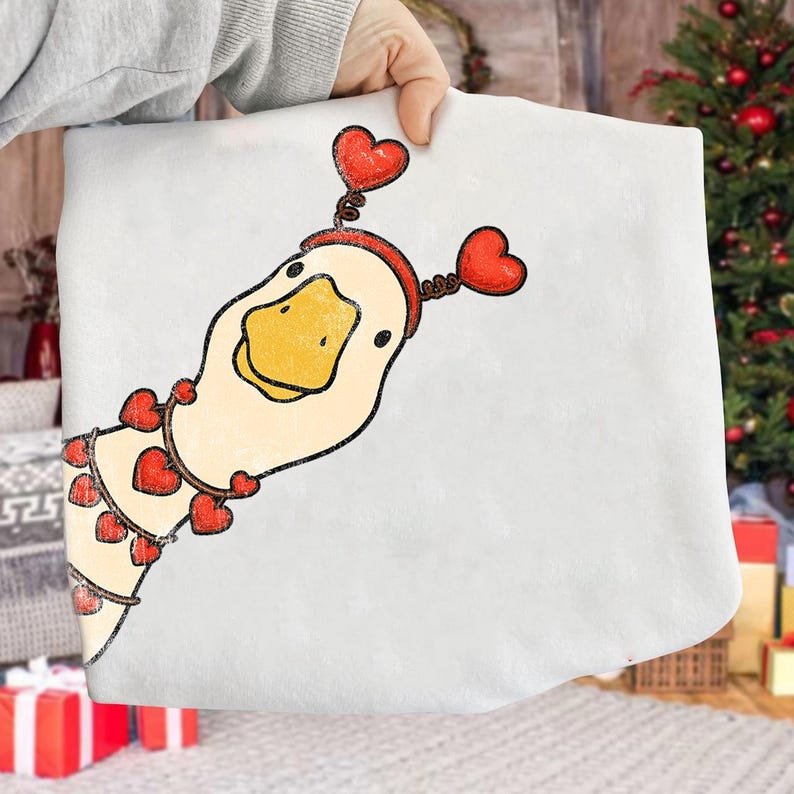 May include: A white, rectangular item featuring a cartoon goose with a yellow beak and a red heart-shaped headband. The goose has a garland of red hearts around its neck. The item is being held up by a person.