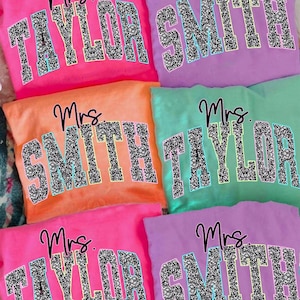 May include: Six colorful sweatshirts in neon pink, lavender, orange, and mint green. Each features the text Mrs. in black script above block letters reading Taylor or Smith. The letters have a black and white speckled pattern with a light blue outline.