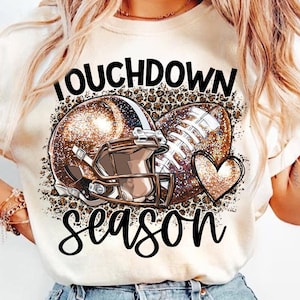 Puede incluir: Una camiseta blanca con un casco de fútbol americano marrón y un gráfico de fútbol. El gráfico incluye un corazón y el texto "Touchdown Season".