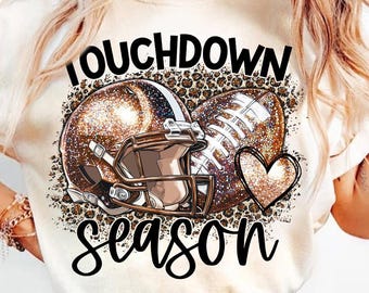 May include: A white t-shirt with a brown football helmet and football graphic. The graphic includes a heart and the text "Touchdown Season".