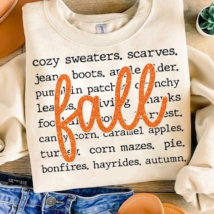May include: A white sweatshirt with the word "fall" in orange glitter lettering on top of a list of fall-themed words. The words include "cozy sweaters", "scarves", "jean boots", "pumpkin patch", "leaves", "food", "bonfires", "hayrides", and "autumn".
