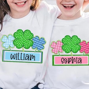 May include: Two white t-shirts with green four-leaf clover designs. The shirt on the left has a blue and white checkered clover, a green clover with white dots, and a blue and white chevron clover. The shirt on the right has a pink and white checkered clover, a green clover with white dots, and a pink and white chevron clover. The shirt on the left has the name "William" printed on a blue banner. The shirt on the right has the name "Sophia" printed on a pink banner.