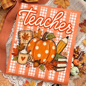 May include: An orange and white gingham patterned t-shirt featuring the word Teacher in orange script. The design includes a large orange pumpkin with white polka dots, a mug of coffee, a pencil, a spiral notebook, and stacked books.