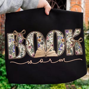 K&ouml;nnte beinhalten: Schwarze Tragetasche mit den W&ouml;rtern "BOOK" und "WORM". Das Wort "BOOK" ist mit einem floralen Muster gef&uuml;llt, und der Buchstabe "O" hat ein offenes Buch. Braune Schleifen schm&uuml;cken die Buchstaben "B" und "K". Das Wort "WORM" ist kursiv geschrieben.