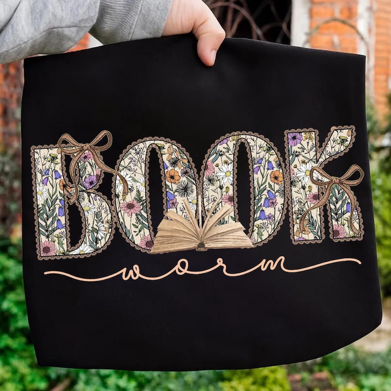 K&ouml;nnte beinhalten: Schwarze Tragetasche mit den W&ouml;rtern "BOOK" und "WORM". Das Wort "BOOK" ist mit einem floralen Muster gef&uuml;llt, und der Buchstabe "O" hat ein offenes Buch. Braune Schleifen schm&uuml;cken die Buchstaben "B" und "K". Das Wort "WORM" ist kursiv geschrieben.