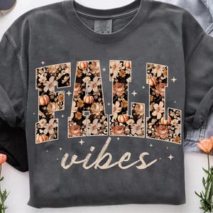May include: Dark gray sweatshirt with the words "FALL vibes" in a floral design with pumpkins. The letters are filled with a pattern of flowers and pumpkins in shades of brown, orange, and cream.
