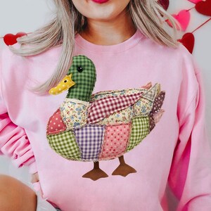 May include: A light pink sweatshirt featuring a patchwork duck design. The duck has a green checkered head, a yellow beak, and a body composed of various fabric patterns, including floral, polka dot, and checkered designs. The duck's feet are brown.