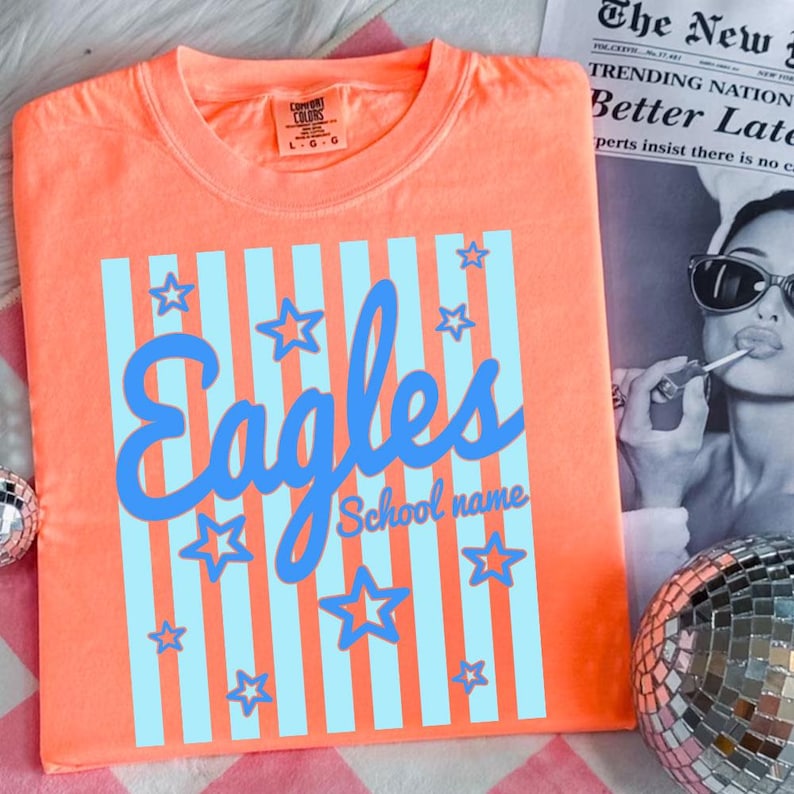 Puede incluir: Una camiseta de color coral con un dise&ntilde;o de rayas azules y blancas. La palabra "Eagles" est&aacute; escrita en cursiva, con estrellas y las palabras "School name" en una fuente m&aacute;s peque&ntilde;a. La camiseta tiene una etiqueta que dice "Comfort Colors".