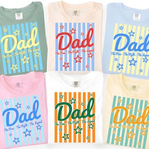 Könnte beinhalten: Sechs T-Shirts in verschiedenen Farben, jedes mit dem Wort "Dad" in einer stilisierten Schriftart und dem Satz "The Man - The Myth - The Legend" darunter. Die Designs umfassen Sterne und gestreifte Hintergründe.