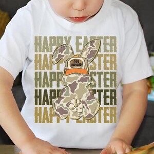 May include: White t-shirt with a camouflage Easter bunny wearing a hat that says "Egg Hunter". The words "Happy Easter" are repeated in gold glitter lettering.