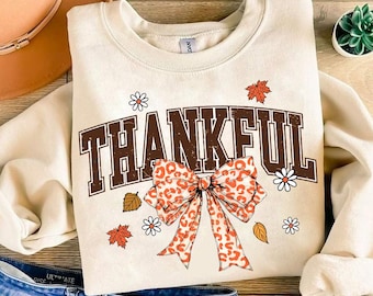 May include: A beige sweatshirt with the word "THANKFUL" printed in brown letters. A large orange and white bow with a leopard print is centered below the word. The bow is surrounded by small white flowers and orange leaves.