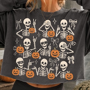 May include: A charcoal gray long-sleeve sweatshirt featuring a repeating graphic print of white skeletons and orange jack-o'-lantern pumpkins. The skeletons are depicted in various poses with bows and star accents scattered across the fabric.